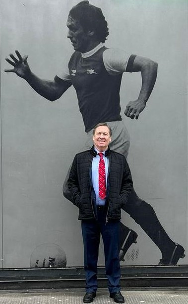 Kenny Sansom in a suit standing outside the Emirates Stadium in front of a large photo of himself